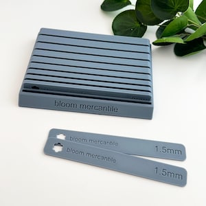 May include: A grey plastic organiser with 10 slots for storing items. The organiser has the text "bloom mercantile" printed on the front. Two grey plastic measuring sticks with the text "bloom mercantile" and "1.5mm" printed on them.