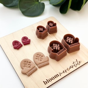 May include: A wooden cutting board with the text "bloom mercantile" features several brown and maroon mitten-shaped cookie cutters. The cutters have snowflake and ribbed designs. Green leaves are visible in the background.