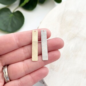 May include: Two rectangular metal earring charms, one gold-toned and one silver-toned, each with a textured surface and a small hole at the top. The charms are displayed against a neutral background, suggesting a jewelry-making theme.