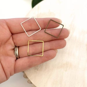 May include: Three square-shaped metal jewelry findings in silver, gold, and bronze tones. The open-frame squares are held in a hand, showcasing their different finishes. The findings are likely used for jewelry making or crafting.