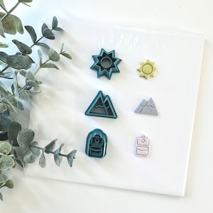 Op de afbeelding: Een set van zes koekjesvormen in verschillende vormen, waaronder een zon, bergen, een rugzak en een ster. De vormen zijn gemaakt van groen, geel en grijs plastic.