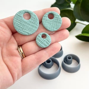 May include: Three teal green textured round polymer clay earrings with a hole in the center. The earrings are arranged in order of size from largest to smallest. The earrings are on a hand with a gold ring.