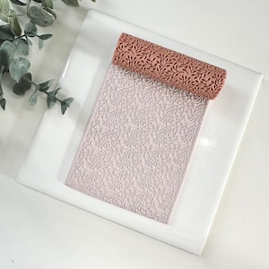 May include: A brown rolling pin with a leafy pattern, resting on a white surface with a matching embossed pattern. The rolling pin is used for creating decorative patterns in fondant or clay.