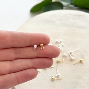 May include: A close-up of several small stud earrings. Two earrings are held in a hand, one silver and one gold. Other earrings are scattered on a light-colored surface. The earrings have a small ball at the end of a post.