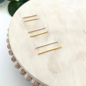 May include: An assortment of small, straight metal bars in silver and gold tones. The bars are arranged on a light-colored wooden surface. Each bar has a small hole at each end, suggesting they are components for jewelry making or crafts.