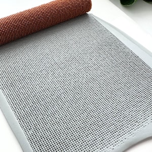 May include: A rolled brown textured roller sits atop a gray, grid-patterned mat. The mat has a light gray border. The items are on a white surface with green leaves in the background. This image is suitable for craft projects.