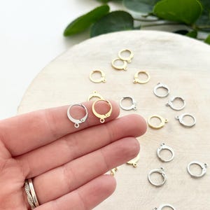 May include: A collection of earring clasps in gold and silver tones. The clasps are small, circular, and have a hinged design for securing earrings. Some clasps are held in a hand, while others are scattered on a light-colored surface.