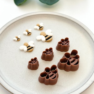 May include: A set of brown and white bee-shaped cookie cutters and fondant toppers. The cutters are in the shape of a bee with wings and a honeycomb pattern. The toppers are in the shape of a bee with wings and a black and yellow striped body.