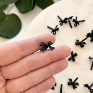 May include: A close-up of a hand holding a black bow. The bow is small and delicate, and it is made of a smooth, shiny material. There are several other black bows scattered on a wooden surface in the background.