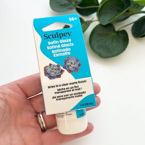 Puede incluir: Un frasco de 30 ml de Sculpey Satin Glaze con una tarjeta de cartón azul y blanca. El empaque presenta el texto Sculpey Satin Glaze, Satiné Glacis, Satinado Esmalte y Dries to a clear matte finish junto a imágenes de dos flores pequeñas.