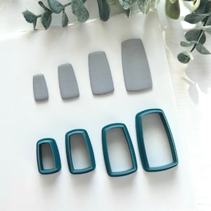 May include: A set of five rectangular polymer clay cutters in various sizes. The cutters are teal green and are arranged in a row on a white surface. Next to the cutters are five rectangular pieces of gray polymer clay, also arranged in a row.