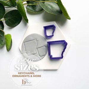 May include: Two purple plastic cookie cutters in the shape of the state of Arkansas. The cutters are on a white hexagonal surface. The text "new sizes, keychains, ornaments & more" is printed below the cutters.