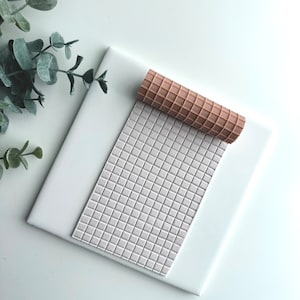 May include: A light brown textured rolling pin rests on a gray, grid-patterned mat, atop a white surface. The rolling pin has a waffle-like design. Greenery is visible in the upper left corner. The items are likely for crafting or baking.