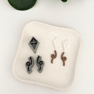 Könnte beinhalten: Ein Set aus vier Ohrringen aus Polymer Clay in verschiedenen Formen und Farben. Die Ohrringe umfassen eine graue Diamantform, zwei graue gebogene Formen und zwei weiße Papierfliegerformen mit braunen Stielen.