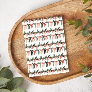 May include: A rectangular greeting card with a festive pattern of red, green, and pink bows and evergreen branches on a white background. The card is displayed on a wooden tray, suggesting a holiday theme.