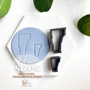 May include: Two black plastic cookie cutters in the shape of the state of Vermont. The cutters are on a white surface with a blue clay impression of the state of Vermont in the background. The text "new sizes" is printed on the surface. The text "keychains, ornaments & more" is printed below the text "new sizes".