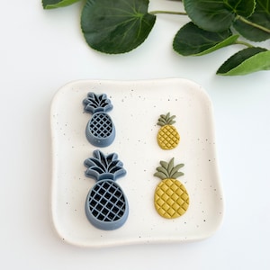May include: Four pineapple shaped cookie cutters, two are blue and two are yellow. The cookie cutters are on a white speckled plate.