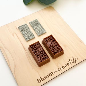 May include: A wooden cutting board with the text "bloom mercantile" is the base for four rectangular clay cutters. Two are gray, and two are brown. The cutters have a floral and geometric pattern. The board is light brown.