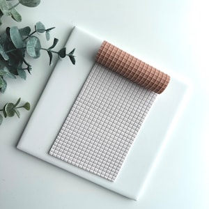 May include: A white square tile with a light grey textured surface. A light brown waffle-like roller is partially rolled up on the tile.