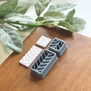 May include: A set of three gray and white plastic cookie cutters shaped like a chevron pattern. The cutters are on a wooden surface.