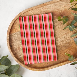 May include: A rectangular notepad with a vertical striped pattern in shades of red, pink, and green. The notepad is resting on a wooden tray with a light-colored background and green foliage.