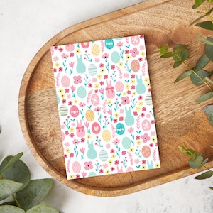 Pode incluir: Um cartão branco com um padrão repetido de coelhos cor-de-rosa, ovos amarelos e ovos azuis com flores cor-de-rosa e folhas verdes.
