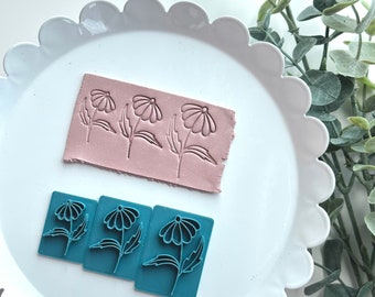 May include: Three blue polymer clay stamps with a daisy flower design. The stamps are on a white plate with a scalloped edge. A pink polymer clay sheet with three daisy flower designs is in the background.