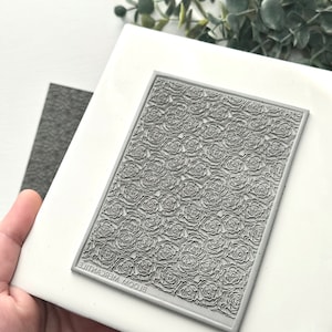May include: A gray rectangular stamp with a rose pattern is held over a white tile. The stamp has the text "BLOOM MERCANTILE" printed in reverse along the bottom edge. A gray felt square is partially visible.