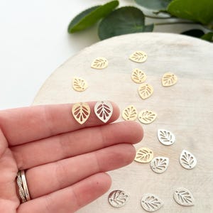 May include: Close-up of leaf-shaped charms in gold and silver tones. Two gold charms are held in a hand, with several more scattered on a wooden surface. The charms have a cut-out leaf vein design, perfect for jewelry making.
