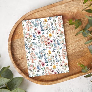 以下が含まれることがあります： ピンク、ブルー、イエロー、ブラウンの花が描かれた花柄の白いカード。花は白い背景に繰り返しパターンで配置されています。