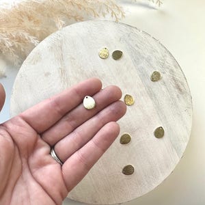 Op de afbeelding: Kleine, goudkleurige metalen bedels in de vorm van een druppel met een getextureerd oppervlak. Sommige bedels hebben een klein gaatje aan de bovenkant. De bedels worden getoond op een wit, rond houten oppervlak.