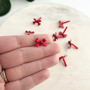 Pode incluir: Pequenos pingentes de laço vermelho espalhados em uma superfície de madeira. Um laço é segurado em uma mão, mostrando os detalhes delicados. Estes laços em miniatura são ideais para artesanato ou joalheria.