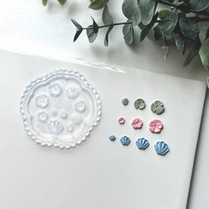 May include: A clear silicone mold with a scalloped edge and eight different flower and shell designs. The mold is shown with several cured resin pieces in green, pink, and blue.