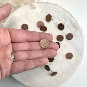 May include: A hand holding several small, round, wooden craft pieces with a hole at the top. The pieces are various shades of brown and are scattered on a white surface.