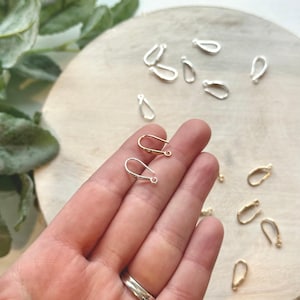 May include: A close-up of a hand holding two silver earring hooks. The hooks are in the shape of a loop with a small hole at the top. There are several other earring hooks in the background, some silver and some gold.