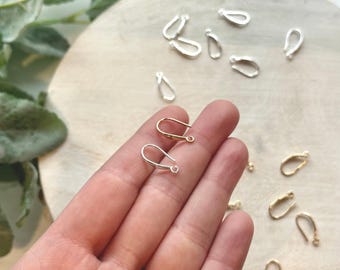 May include: A close-up of a hand holding two silver earring hooks. The hooks are in the shape of a loop with a small hole at the top. There are several other earring hooks in the background, some silver and some gold.