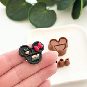 May include: A black polymer clay cutter in the shape of a mouse head with a red bow, a plant, and books. Additional brown cutters are also visible.