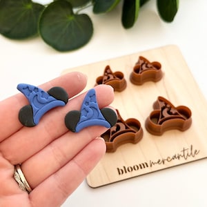 Op de afbeelding: Blauwe en zwarte koekjessnijders in de vorm van een tovenaars hoed met ster- en maandetails. Bruine koekjessnijders in dezelfde vorm liggen op een houten plank met de tekst "bloom mercantile".