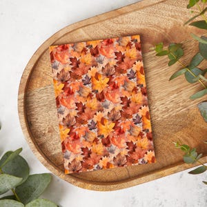 Puede incluir: Un cuaderno con un diseño de acuarela de hojas de otoño en tonos naranja, rojo y marrón. Las hojas están dispuestas en un patrón repetitivo. El cuaderno está sobre una bandeja de madera con hojas de eucalipto.