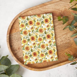 Könnte beinhalten: Eine weiße Karte mit einem sich wiederholenden Muster aus gelben Sonnenblumen und grünen Blättern. Die Karte liegt auf einem Holztablett.