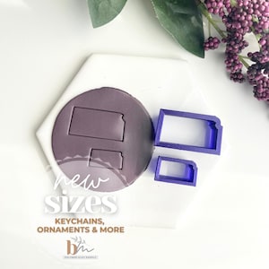 Peut inclure: Deux emporte-pièces en plastique violet en forme de l'état du Kansas. Les emporte-pièces sont sur une surface blanche avec le texte "new sizes, keychains, ornaments & more" imprimé en dessous.