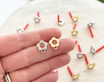 May include: Close-up of flower-shaped earring studs in silver and gold tones. The earrings have a small loop at the bottom and are displayed on a wooden surface. Red earring posts are visible in the background.