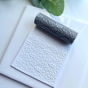 May include: A grey textured rolling pin with a decorative pattern. The rolling pin is on a white surface and has been used to create an embossed pattern on the surface.