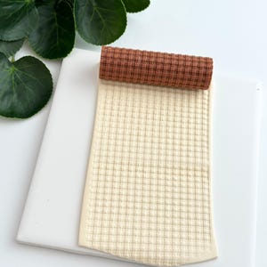 May include: A rolled brown and cream-colored fabric with a checkered pattern sits atop a white square surface. The fabric is a waffle weave, and the roll is a solid brown color. Green leaves are visible in the background.