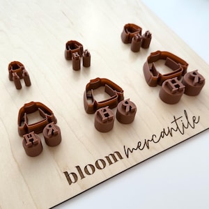 May include: A selection of brown plastic cookie cutters and stamps on a light wooden board. The cutters are shirt-shaped, and the stamps feature the letters "H" and "R". The board is engraved with the text "bloom mercantile".