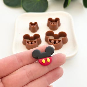May include: A selection of brown plastic cookie cutters shaped like a cartoon mouse head, alongside a matching decorative charm. The charm is black, red, and yellow. The cutters are on a white plate, with green leaves in the background.