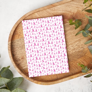 May include: A greeting card with a white background covered in pink awareness ribbons. The card is on a wooden tray with a light-colored wood grain. Green leaves are in the background.