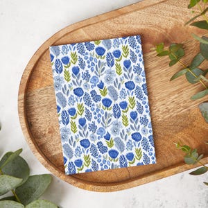 Puede incluir: Una tarjeta rectangular con un estampado floral con flores azules y celeste y hojas verdes sobre fondo blanco. La tarjeta está colocada sobre una bandeja de madera con acabado marrón claro. Hojas de eucalipto en el fondo.