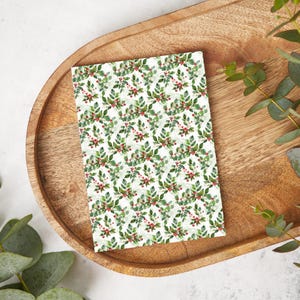 May include: A rectangular card with a repeating pattern of green leaves and red berries on a white background. The card is resting on a wooden tray, with green foliage in the background. The design evokes a festive, holiday theme.
