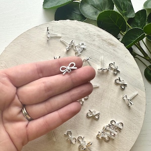 May include: Silver bow charms with clear plastic earring backs. The charms are small and delicate, perfect for adding a touch of elegance to jewelry.
